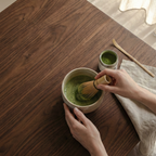 Person preparing matcha in a small bowl on a wooden table with a soft light background