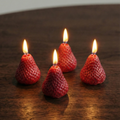 Four strawberry-shaped candles with lit wicks on a wooden surface.